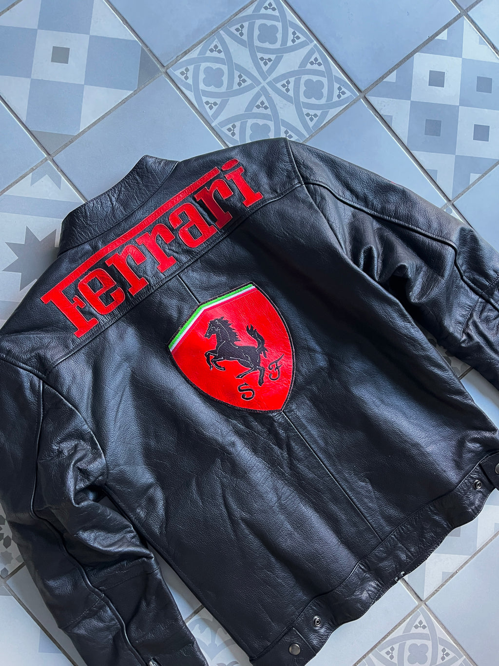 Dos du blouson cuir Ferrari vu de biais, mettant en valeur le grand écusson brodé du Cheval Cabré et le lettrage rouge Ferrari.