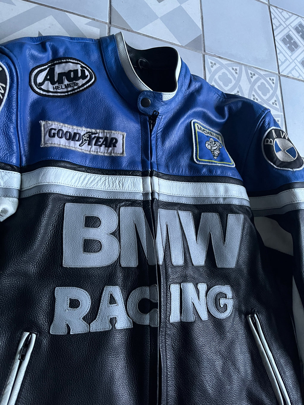 Gros plan sur le col motard et la poitrine du blouson cuir BMW, montrant les écussons brodés Arai, Goodyear, Michelin et la texture du cuir patiné.